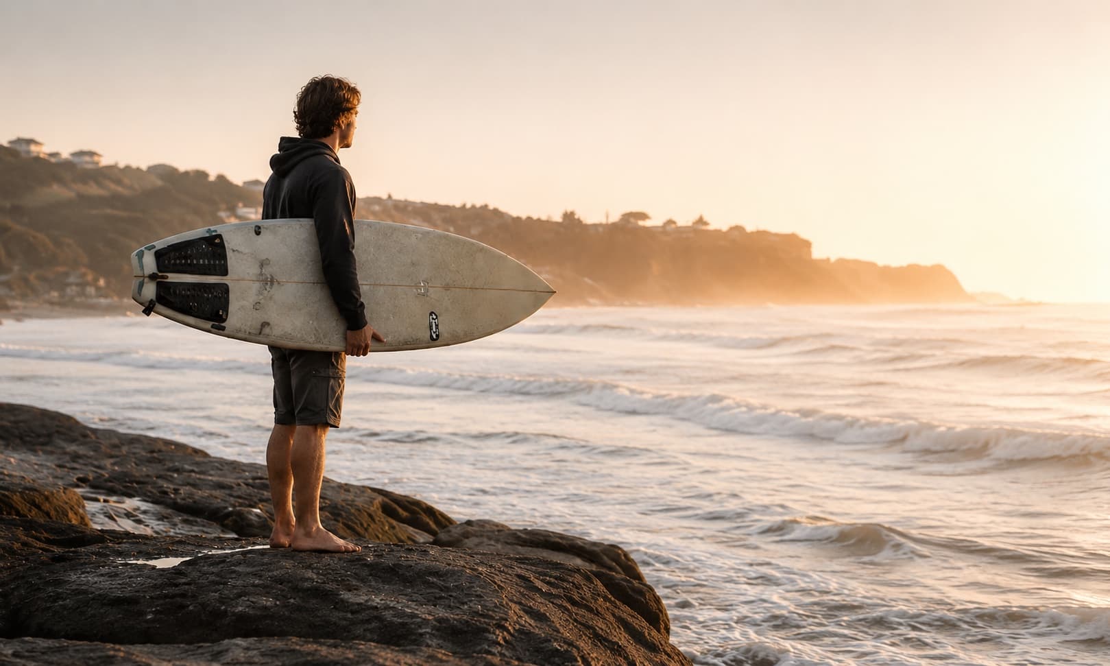 Nathan standing on a beach at sunrise holding a surfboard, looking out at the ocean